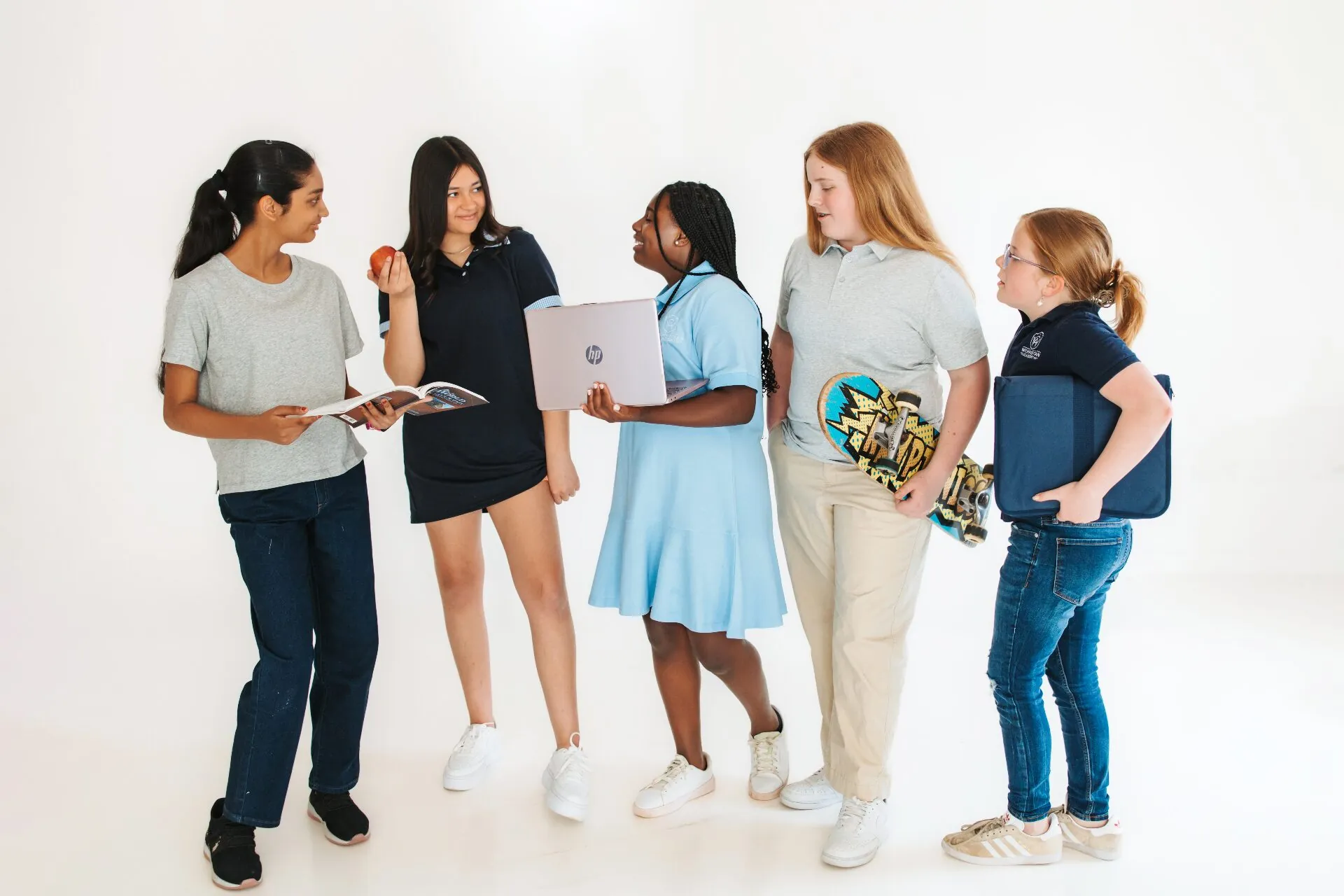 A diverse group of Way Christian Academy students standing together with books, a laptop, a skateboard, and a folder in a bright studio