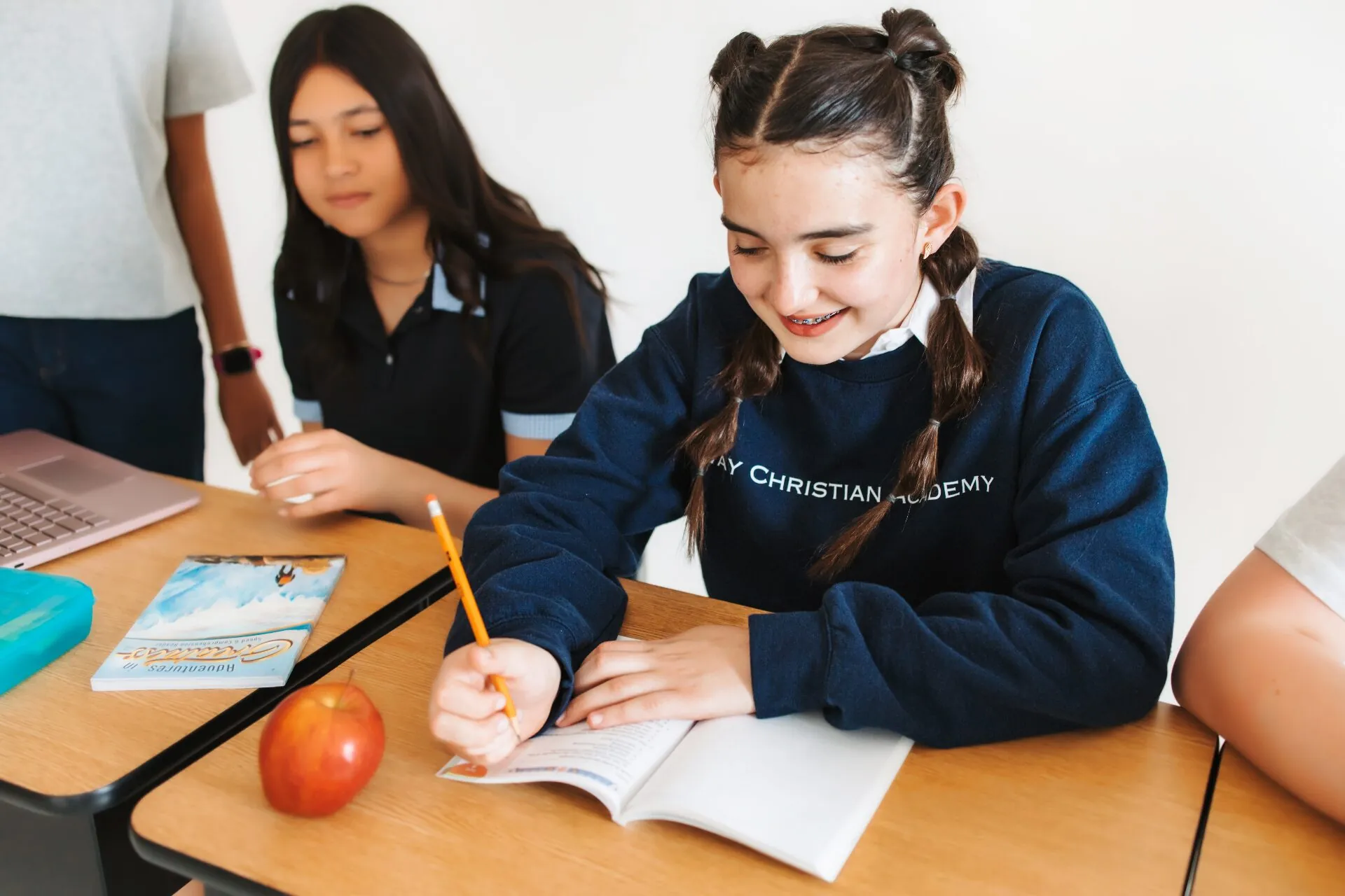 A Way Christian Academy student smiling while writing in a workbook at her desk, with a classmate studying nearby