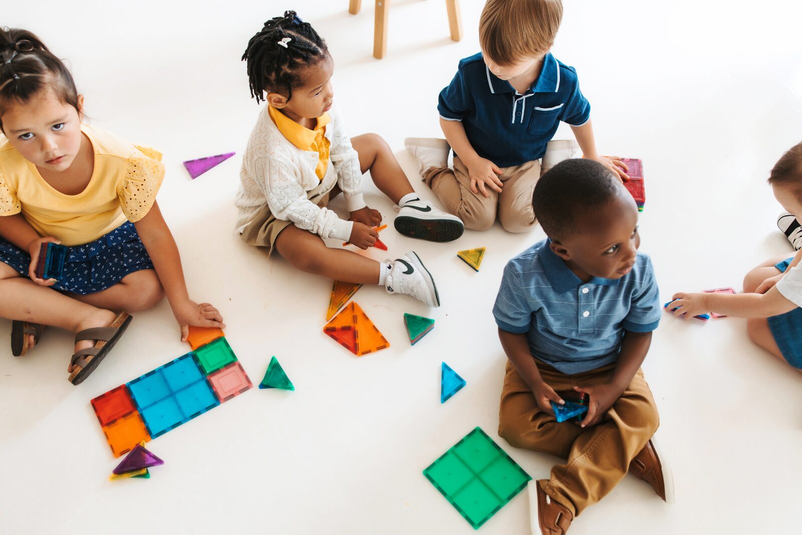 Young children sitting on the floor playing with colorful magnetic tiles in a bright, cheerful classroom